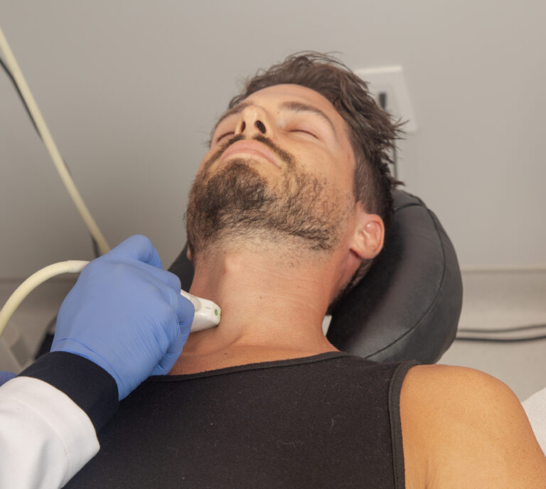 A doctor wearing blue latex gloves, holds an Ultrasound scanner against the neck of a male patient before administering a Stellate Ganglion Block injection into the patient's neck.