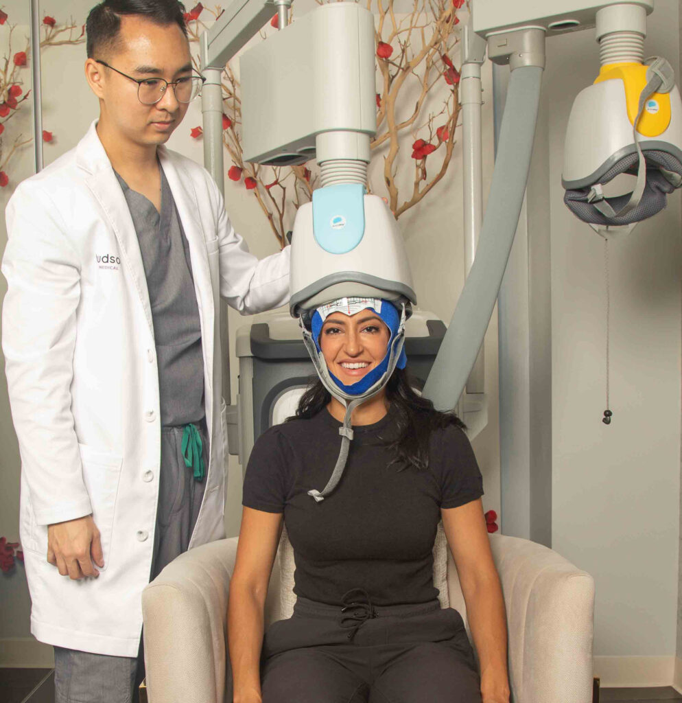 A woman sits under a Transcranial Magnetic Stimulation (TMS) helmet while a male Physician Assistant stands beside her and monitors the magnetic pulses emitted by the TMS machine.