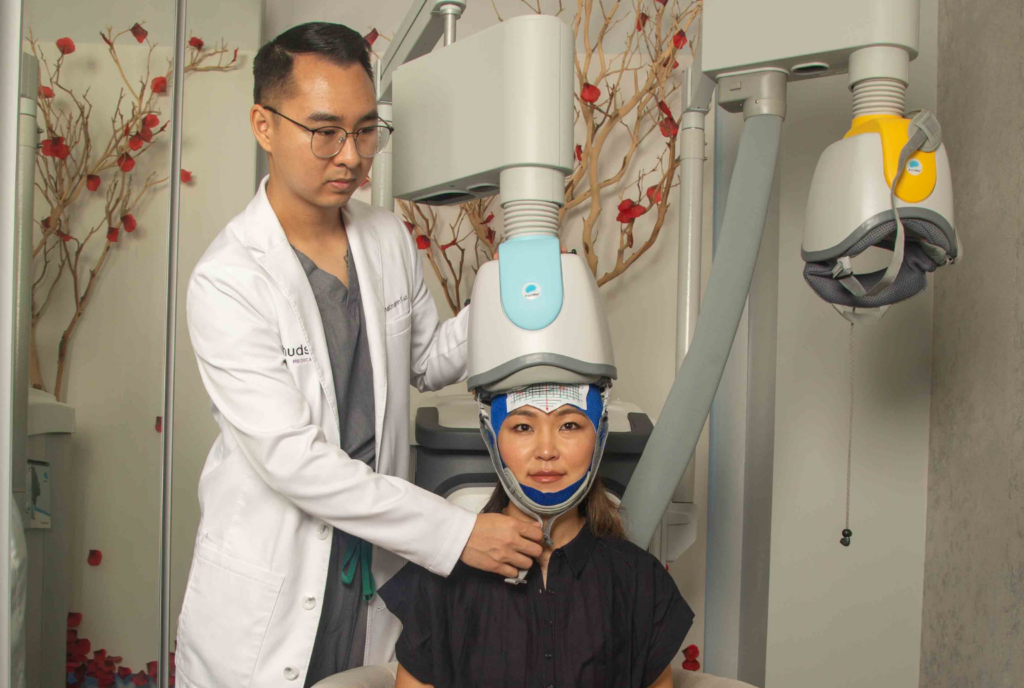 male physician from Hudson Mind fitting a Transcranial Magnetic Stimulation (TMS) helmet on the head of a female patient.