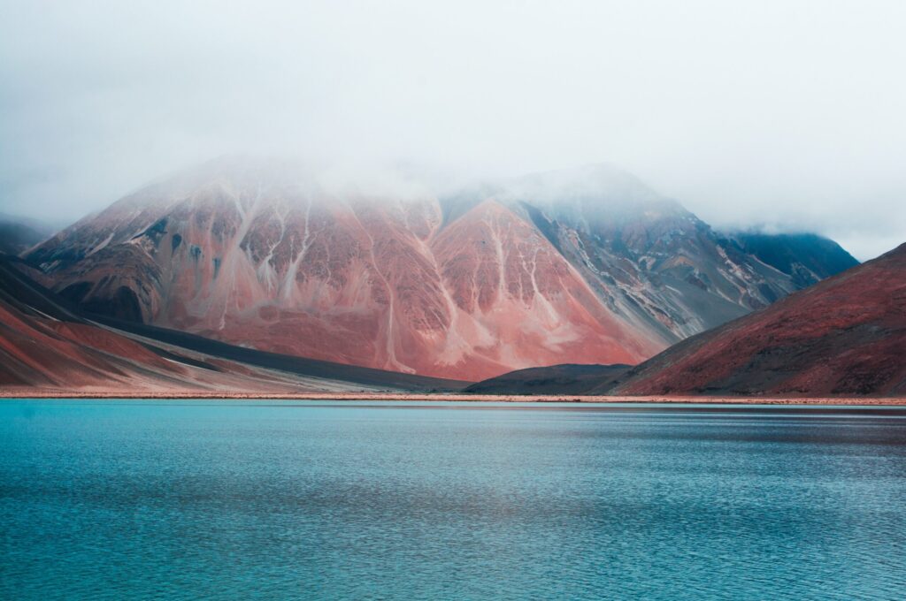 A calm, blue lake in front of a red-tinted mountain, whose with peak is hidden behind clouds