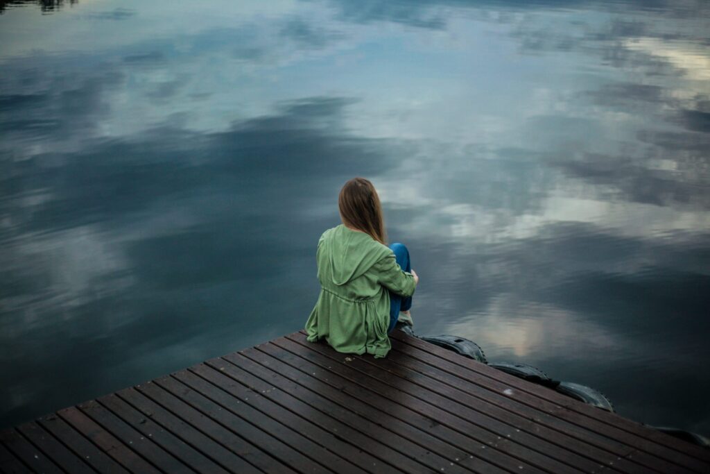 a woman wearing a green raincoat sits on the ledge of a dock staring down at the surface of a lake that reflects a cloudy sky.